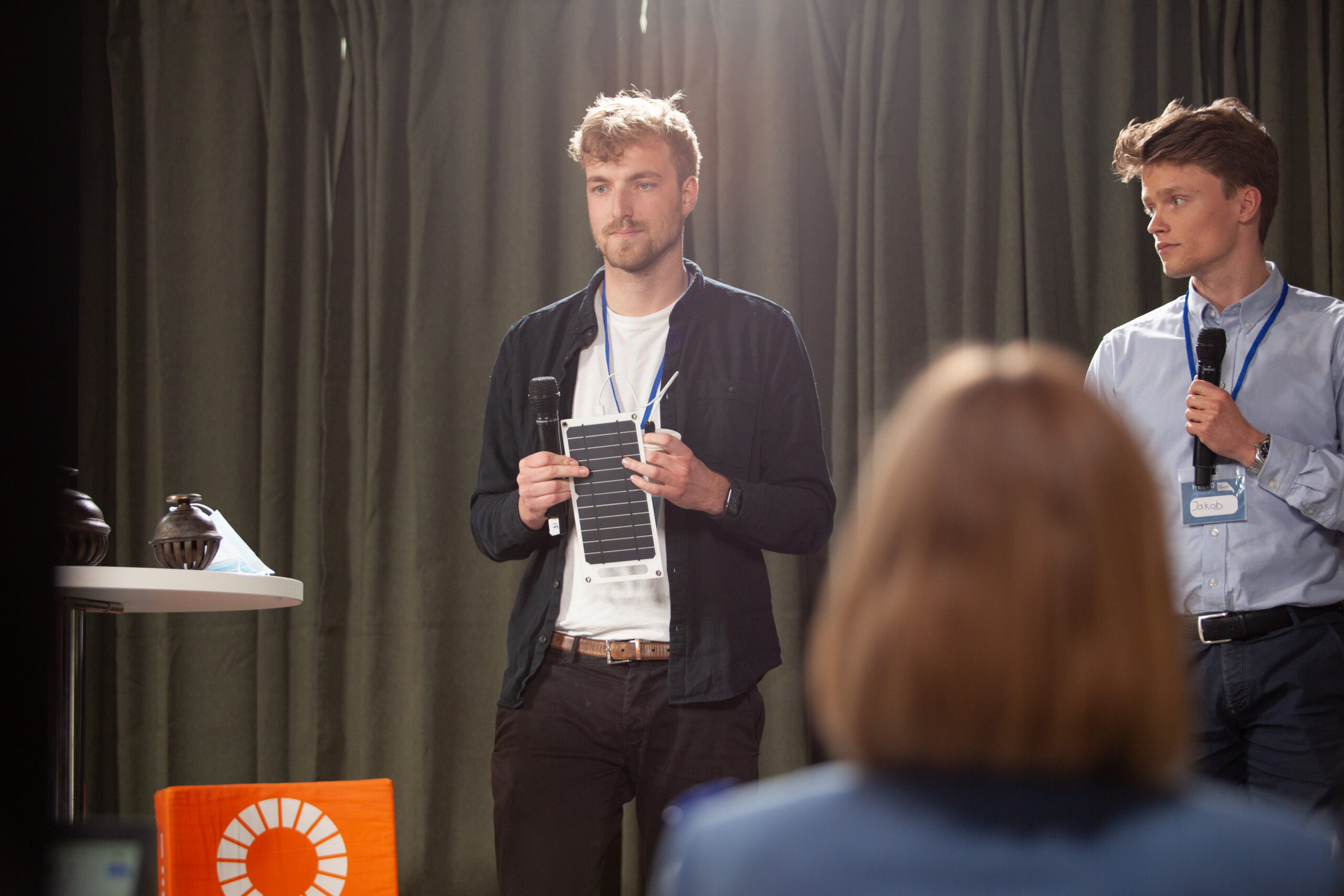 Two Male students pitching their bold idea on stage. One is holding a part of a prototype, the other a microphone